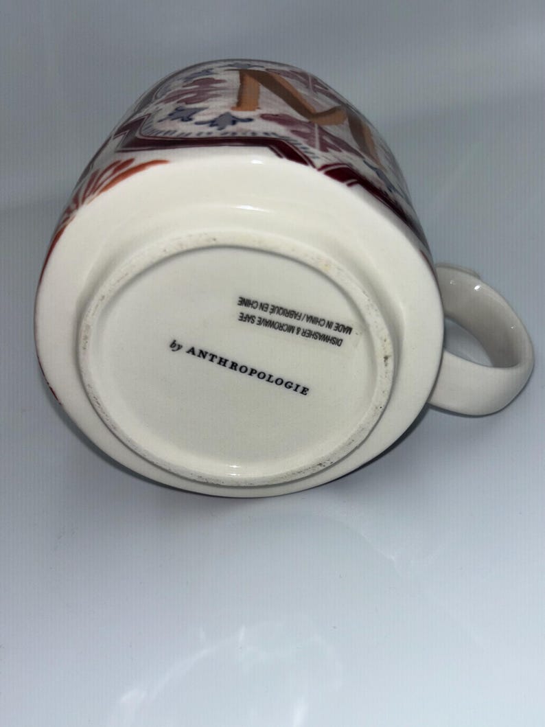 Op de afbeelding: Witte keramische mok met een decoratief patroon in bruin-, rood- en blauwtinten. De mok heeft een oor en de tekst "by ANTHROPOLOGIE" is onderaan zichtbaar. De mok is vaatwasser- en magnetronbestendig.