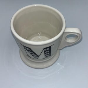 Op de afbeelding: Cr&egrave;mekleurige keramische mok met een zwart letter "M" ontwerp. De mok heeft een afgeronde basis en een handvat. De letter "M" is gestileerd met zwarte lijnen. De mok is leeg en staat op een wit oppervlak.