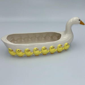 May include: A white ceramic serving dish shaped like a long goose with an elongated neck and orange beak. The exterior of the dish is decorated with a row of eight small, bright yellow ducklings attached along the side of the oval-shaped bowl.