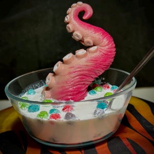 May include: A clear glass bowl filled with milk and colorful cereal, with a pink and tan octopus tentacle rising from the cereal. A spoon rests in the bowl. The background is dark.