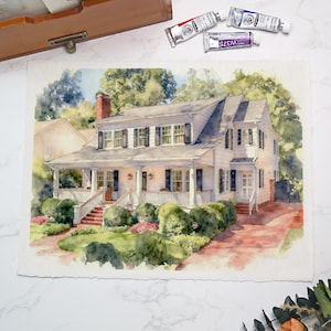 May include: Watercolor painting of a two-story white house with a porch, black shutters, and a red brick chimney. The house is surrounded by green bushes and trees. Paint tubes are visible in the top right corner.