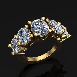 Könnte beinhalten: Ein goldener Ring mit fünf oval geschliffenen Diamanten. Die Diamanten sind in einer Reihe angeordnet, wobei der größte Diamant in der Mitte liegt. Der Ring hat eine polierte Oberfläche und einen schwarzen Hintergrund.