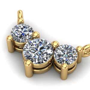 Pode incluir: Close-up de um pingente de ouro com três pedras preciosas redondas e transparentes. As pedras são fixadas em garras de ouro e dispostas em linha curva. O pingente tem uma pequena argola em cada extremidade para uma corrente.