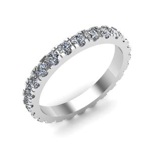 May include: A silver band ring featuring a continuous line of small, clear gemstones. The ring is set against a white backdrop, highlighting its polished metal and sparkling stones. The ring is a classic design.