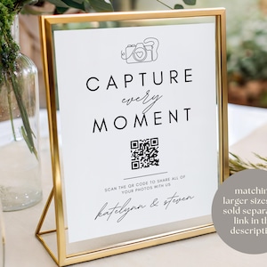 Puede incluir: Un letrero con marco dorado con las palabras "CAPTURE every MOMENT" en escritura negra. Un c&oacute;digo QR est&aacute; debajo del texto, con los nombres "katelynn & steven" en la parte inferior. El letrero est&aacute; sobre una mesa con un fondo borroso.