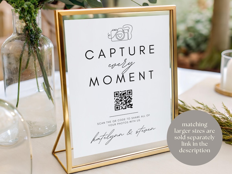 Puede incluir: Un letrero con marco dorado con las palabras "CAPTURE every MOMENT" en escritura negra. Un c&oacute;digo QR est&aacute; debajo del texto, con los nombres "katelynn & steven" en la parte inferior. El letrero est&aacute; sobre una mesa con un fondo borroso.