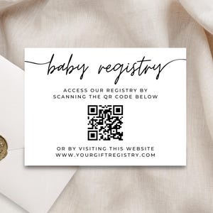 May include: A white baby registry card with black text and a QR code. The card reads "baby registry" in cursive, with instructions to scan the QR code or visit a website. An envelope with a gold seal is in the background.