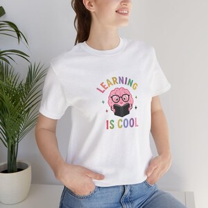 Puede incluir: Una camiseta blanca de manga corta con un gráfico de un cerebro rosa que lleva gafas negras y lee un libro negro. El texto "LEARNING IS COOL" está impreso en letras coloridas encima y debajo de la ilustración del cerebro con estrellas.