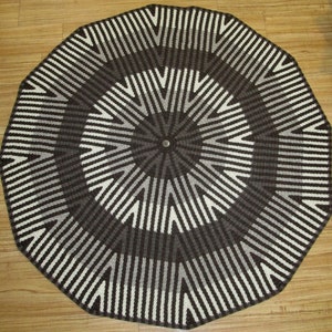May include: A round, crocheted rug with a geometric pattern in shades of brown, white, and gray. The rug has a circular design with a central point and radiating lines.