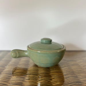 Puede incluir: Un pequeño cuenco de cerámica verde claro para sopa con tapa y asa. El cuenco tiene forma redondeada y un acabado liso y brillante. El asa curvada está unida al lateral. La tapa tiene un pequeño pomo.