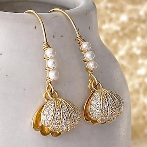 May include: A pair of gold-colored earrings with a seashell design. Each earring features a cluster of small, white pearls and a shell-shaped pendant encrusted with clear crystals. The earrings have a hook closure.