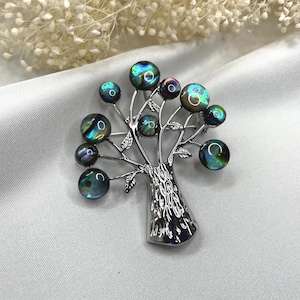 May include: A silver-toned tree brooch with iridescent, round, abalone-like orbs representing the leaves. The brooch has a textured trunk and delicate branches with small, detailed leaves. The orbs display a range of colors, including green, blue, and purple.