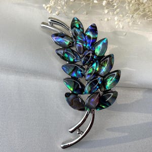 May include: A silver-toned brooch in the shape of a leaf or sprig. The brooch features iridescent, oval-shaped stones in shades of blue, green, and purple. The design has a curved stem and a pair of curved accents at the top.