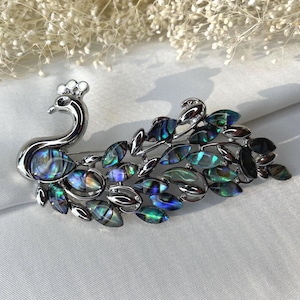 May include: A silver-toned peacock brooch with iridescent abalone shell accents. The brooch features a detailed peacock design with a curved neck, a crown, and a fan of leaf-shaped feathers. The abalone shell provides a colorful, shimmering effect.