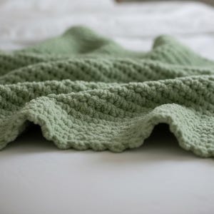May include: A close-up of a soft, sage green blanket with a textured, woven pattern. The blanket is folded and draped, showing the detailed stitching and plush material. The edges have a scalloped design, adding a decorative touch to the cozy home textile.