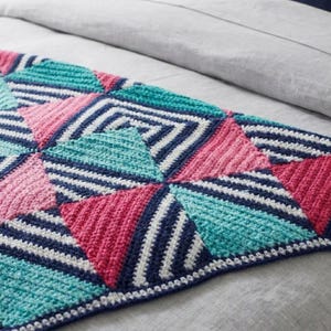 May include: A colorful, geometric crochet blanket featuring a patchwork design. The blanket showcases triangles in shades of pink, teal, and navy blue, with white stripes and a white border. The blanket is displayed on a gray surface.