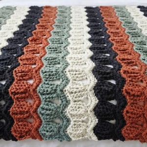 May include: A crocheted blanket featuring a repeating wavy pattern of vertical stripes in alternating colors of cream, sage green, dark charcoal gray, and burnt orange. The texture consists of raised stitches and openwork gaps.