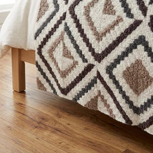 May include: A textured, off-white throw blanket with a geometric diamond pattern in shades of brown, gray, and cream. The blanket is draped over a wooden bed frame, showcasing its soft, plush texture and decorative design.