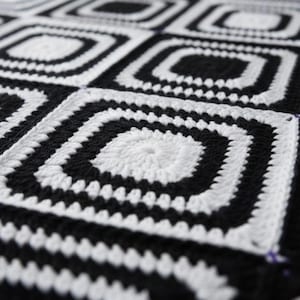 May include: A close-up of a black and white crocheted blanket. The blanket features a repeating pattern of squares with concentric, geometric designs. The color scheme is a stark contrast of black and white, creating a bold visual effect.