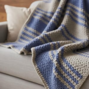 Op de afbeelding: Een blauw en beige gehaakte deken met een streepjespatroon is over een grijze bank gedrapeerd. De textuur van de deken is zichtbaar, met een zacht, handgemaakt uiterlijk. Een wit kussen staat op de achtergrond.