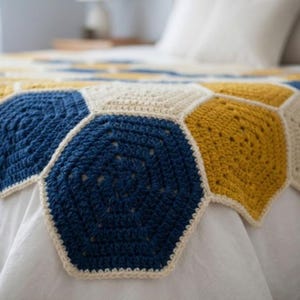 Peut inclure: Une couverture en crochet faite à la main présentant un motif répétitif d'hexagones entrelacés en fil bleu marine profond, jaune moutarde et blanc crème. Le textile affiche une construction en mailles texturées avec des boucles visibles.