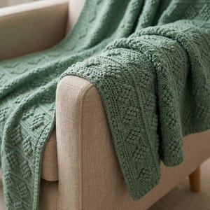 May include: A sage green crochet blanket featuring a textured diamond pattern with raised bobble stitches. The textile is draped over the arm of a piece of furniture, showcasing the intricate, repeating geometric design and the soft, yarn-based construction.