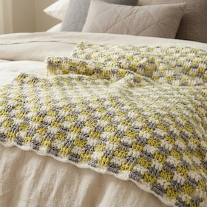 Könnte beinhalten: Eine handgefertigte Häkeldecke mit einem Schachbrettmuster aus Weiß, Grau und Gelbgrün. Die Decke ist über einem Bett drapiert und zeigt ihr strukturiertes Design und ihre weiche Farbpalette. Die Kanten sind mit einem weißen Muschelrand versehen.