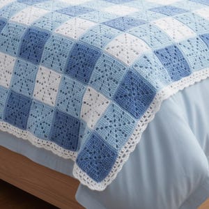 May include: A handmade crochet blanket in shades of blue and white. The blanket features a square pattern with a decorative white border. The blanket is draped over a bed, showcasing its texture and design.