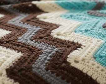 May include: Close-up of a handmade crochet blanket with a chevron pattern. The blanket features brown, cream, grey, and teal blue yarn. The texture of the crochet stitches is visible, creating a cosy and inviting look.
