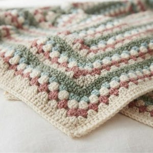 Puede incluir: Una manta de ganchillo con un patrón repetitivo de cuadrados de la abuela en tonos suaves de verde salvia, rosa empolvado, azul claro y crema. El tejido muestra una superficie texturizada con densos grupos de puntos dispuestos en filas.