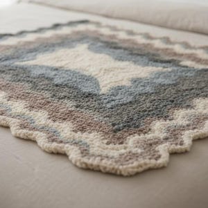 May include: A handmade, textured blanket with a geometric pattern. The blanket features a diamond design in shades of cream, gray, blue, and brown. The edges have a scalloped trim. The blanket is made of soft, plush material.