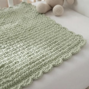 May include: A sage green crochet blanket featuring a textured stitch pattern and a scalloped edge. The blanket is crafted from yarn and displays a consistent, repeating knit design throughout the fabric, finished with a decorative border.
