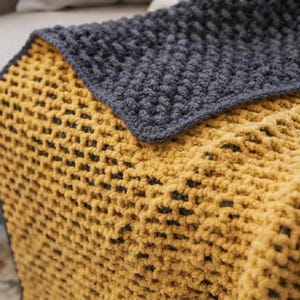 May include: A chunky knit blanket featuring a two-tone design with a mustard yellow side and a dark charcoal gray side. The fabric has a thick, textured waffle-like stitch pattern throughout, showcasing the soft yarn loops and the contrasting colors.