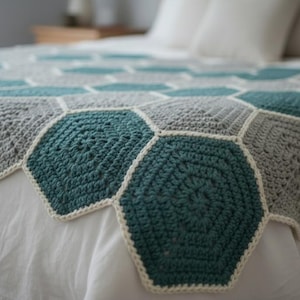 Può includere: Una coperta all'uncinetto fatta a mano con un motivo geometrico di esagoni turchese e grigi. La coperta ha un bordo color crema ed è esposta su un letto bianco. Questa accogliente coperta è perfetta per aggiungere un tocco di fascino fatto a mano a qualsiasi camera da letto.
