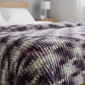 May include: A close-up of a plush, textured blanket in shades of purple, gray, and cream. The blanket is draped over a bed, showcasing its soft, chunky knit design. The color palette creates a cozy and inviting aesthetic, perfect for home decor.