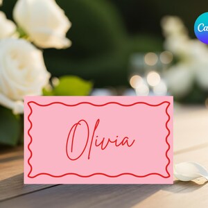 May include: A pink place card with a red wavy border and the name "Olivia" in red script. The card is on a wooden surface, with white flowers and greenery in the background.