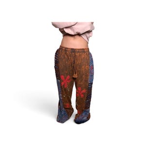 May include: Brown pants with a distressed wash, featuring red floral appliques on the front, blue patterned side panels with a paisley-like design, a drawstring waistband, and vertical pleated detailing along the outer leg seams.