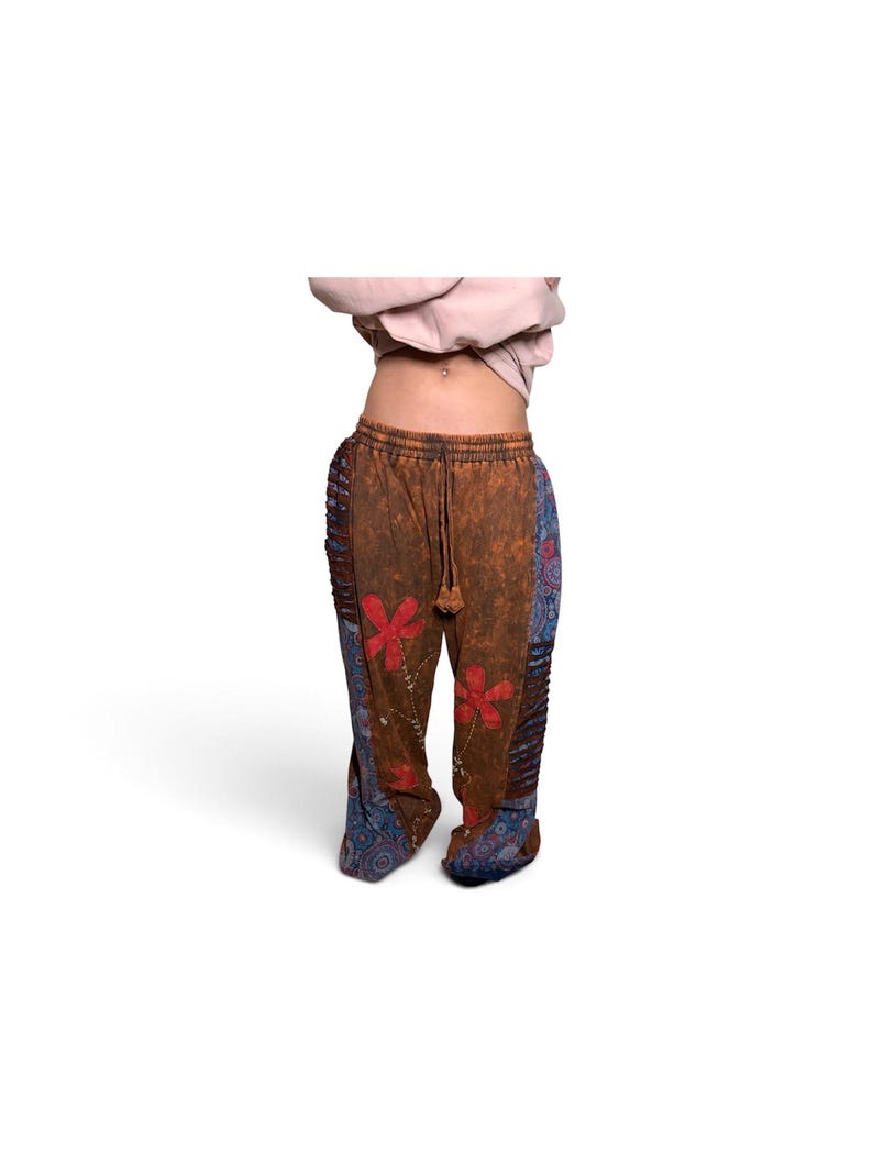 May include: Brown pants with a distressed wash, featuring red floral appliques on the front, blue patterned side panels with a paisley-like design, a drawstring waistband, and vertical pleated detailing along the outer leg seams.