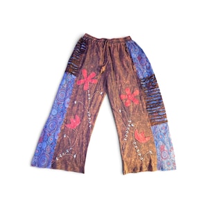 May include: Wide-leg pants featuring a brown acid-wash base with red floral appliqu&eacute;s and embroidered stems. The sides include blue panels with a circular mandala pattern and horizontal brown fabric strips. The waistband has a drawstring closure.