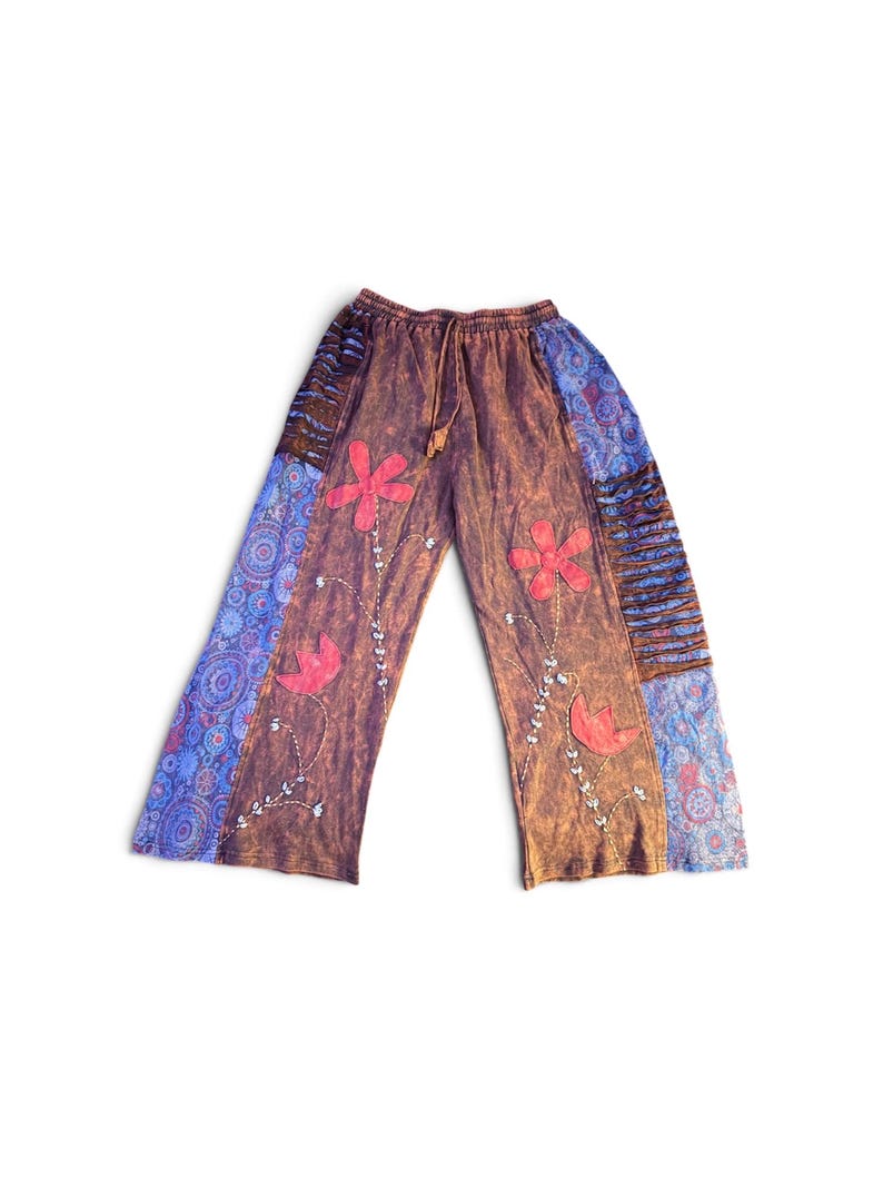 May include: Wide-leg pants featuring a brown acid-wash base with red floral appliqu&eacute;s and embroidered stems. The sides include blue panels with a circular mandala pattern and horizontal brown fabric strips. The waistband has a drawstring closure.