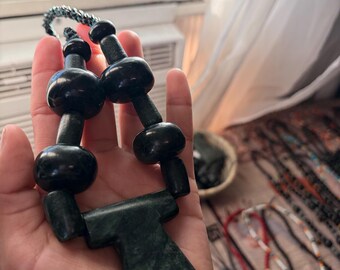 May include: A dark green stone necklace featuring smooth, rounded beads and a large, flat, T-shaped pendant. The necklace is strung on a dark, braided cord, showcasing a deep forest green color with subtle natural marbling throughout the stone pieces.