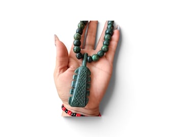 May include: A dark green stone pendant shaped like a stylized paddle with a cross-hatched texture on the front, suspended from a beaded necklace featuring round, mottled dark green stone beads of varying shades and sizes.