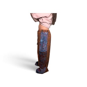 May include: Loose-fitting trousers featuring a brown acid-wash fabric with a vertical panel of blue and red circular mandala patterns. The pants include a drawstring waistband and a section of horizontal cut-out detailing along the side leg.
