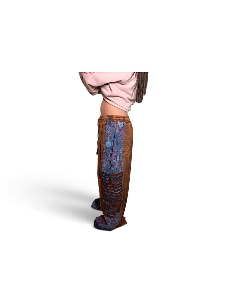 May include: Loose-fitting trousers featuring a brown acid-wash fabric with a vertical panel of blue and red circular mandala patterns. The pants include a drawstring waistband and a section of horizontal cut-out detailing along the side leg.