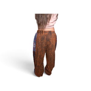 May include: Loose-fitting pants featuring a brown, mottled, acid-wash texture on the front and back panels, contrasted by blue side panels with a repeating circular floral pattern. The pants have an elasticized waistband and a relaxed silhouette.