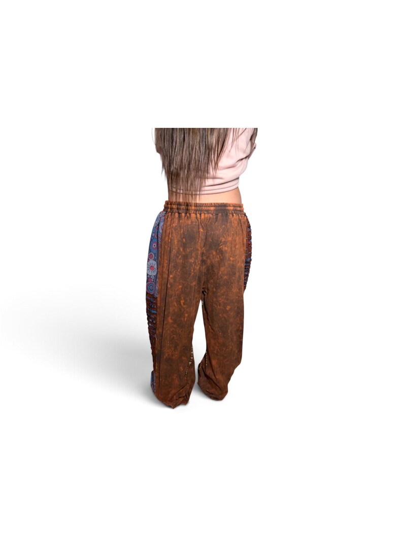May include: Loose-fitting pants featuring a brown, mottled, acid-wash texture on the front and back panels, contrasted by blue side panels with a repeating circular floral pattern. The pants have an elasticized waistband and a relaxed silhouette.