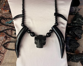 May include: A black necklace featuring a central carved panther head pendant flanked by two large, curved black tusks and smaller black beads. The necklace is strung on a black cord with additional smaller black beads along the upper sections.