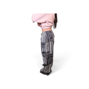 May include: Loose-fitting pants featuring a patchwork design of grey fabric with black and white line illustrations and vertical black and white striped panels. The pants include a side cargo pocket and an elastic waistband with a drawstring closure.