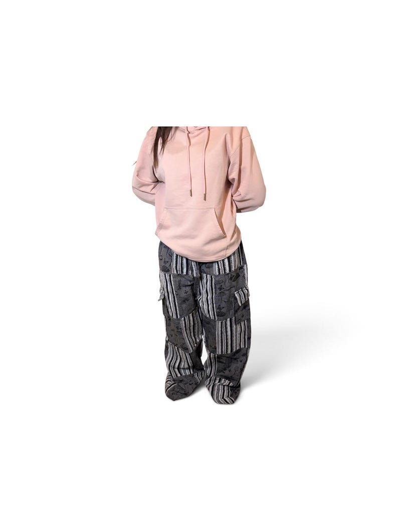 May include: A light pink hooded sweatshirt with a front kangaroo pocket and matching drawstrings, paired with loose-fitting cargo pants featuring a patchwork design of black and gray striped fabric and panels with a dark floral pattern.