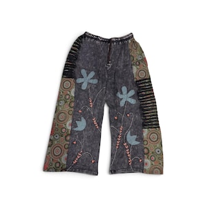 May include: Wide-leg pants in a dark gray acid-wash fabric featuring light blue fabric flower appliqués, embroidered stems with small pink accents, side panels with a multi-colored circular pattern, and black horizontal cut-out detailing.